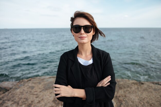 Smiling cute young woman in sunglasses standing with hands folded near the sea