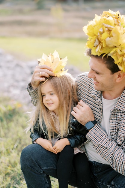 写真 黄色い葉の花輪をかぶった笑顔のお父さんが、座っている小さな女の子の頭に紅葉を置きます