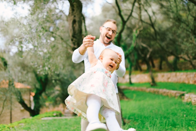 Smiling dad shakes a little laughing girl in his arms in a green park against a background of trees