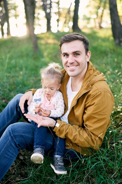 Foto papà sorridente seduto con una bambina sul prato che le mostra una piccola lumaca in un guscio