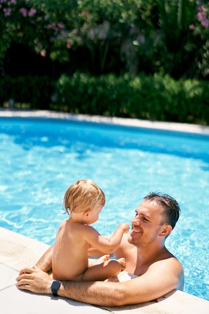 写真 笑顔のパパが水の中に立って ⁇ プールの端に座っている小さな子供を抱きしめています