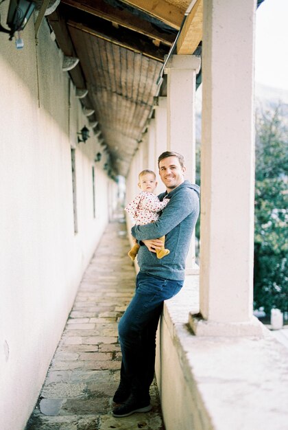 Smiling dad with a baby in his arms stands on the terrace leaning against a stone fence