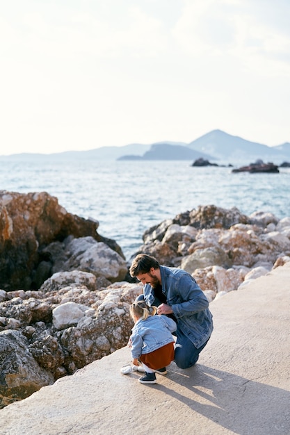 Smiling dad with a little girl squatting on a large rock near the water