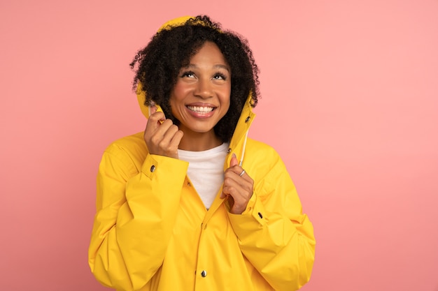 Smiling dark skinned curly woman in yellow raincoat with hood has good mood