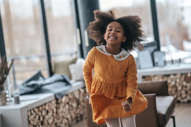 A smiling dark-skinned girl in orange dress dancing in the room