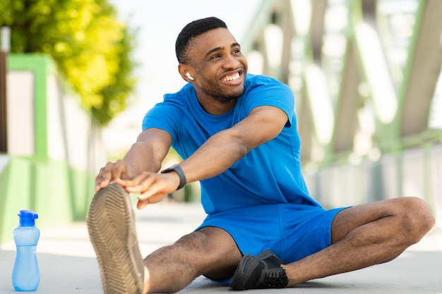 Smiling dark-skinned sportsman sits on a track stretches and listens to music, copy space