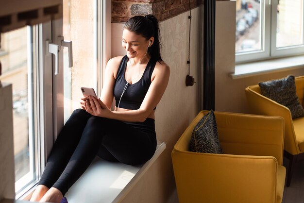 Smiling darkhaired woman in earphones listening music while sitting on windowsill Pretty female in sport clothes using smartphone between exercises at home