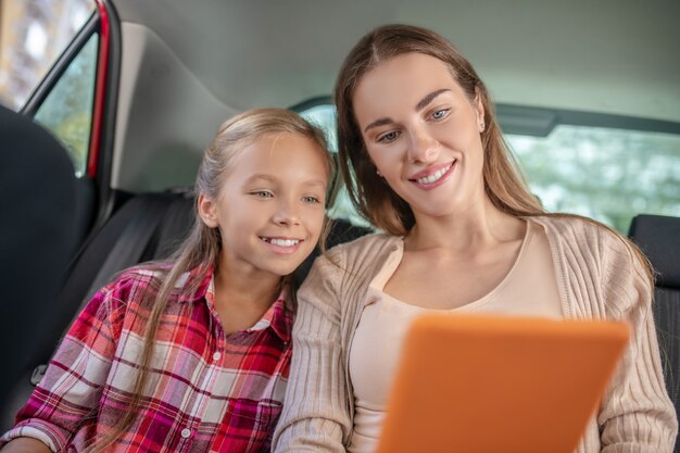 車の後部座席のタブレットで何かをチェックしている笑顔の娘と彼女のお母さん