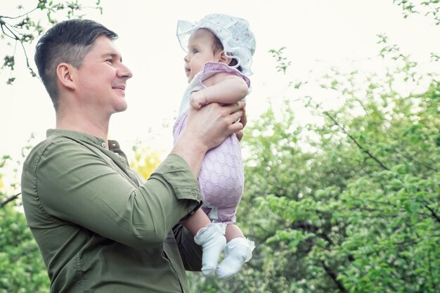 Photo smiling delighted man in shirt holds funny baby girl in arms and tosses standing in green park among blossoming trees on spring day