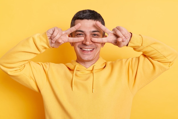 Smiling delighted man wearing casual style hoodie posing isolated over yellow background standing with victory peace sign and looking at camera expressing positive emotions