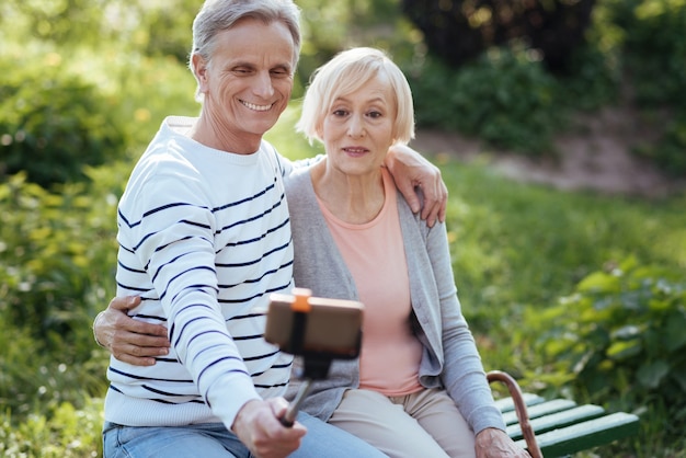 Smiling delighted old couple hugging each other and using selfie stick while enjoying weather in the park and taking pictures