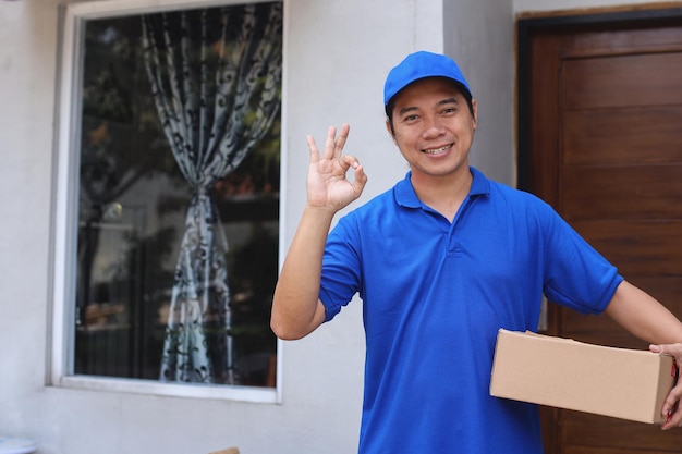 Smiling delivery man in blue uniform with cap holds parcel to send and gesturing ok or good sign