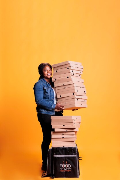 Smiling deliverywoman holding stack full of pizza boxes preparing to deliver to clients during lunch time. Restaurant emloyee with takeout thermal backpack delivering takeaway food order