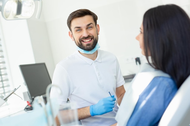 Smiling doctor looking at the camera and holding instruments for looking client sitting in dental chair