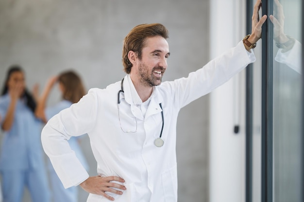 Un medico sorridente in piedi e pensieroso che guarda fuori dalla finestra mentre fa una breve pausa in un corridoio dell'ospedale durante la pandemia di covid-19.