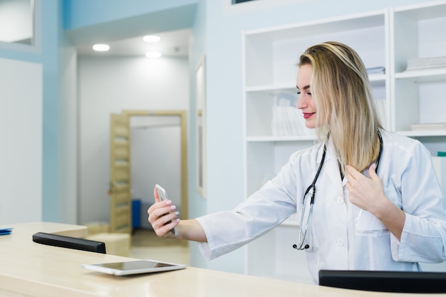 Smiling doctor taking selfies with her phone behind reception desk