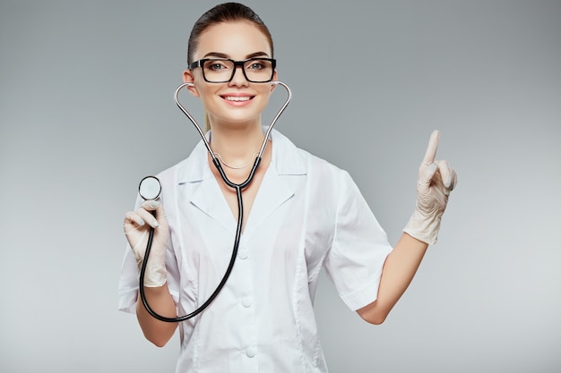 Smiling doctor with brown hair and nude make up wearing white medical uniform, glasses, stethoscopes and white gloves at gray studio background, portrait, pointing with finger.