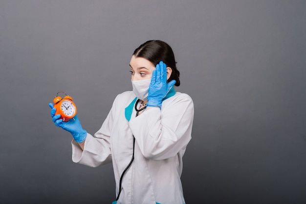 Улыбающаяся женщина-врач в маске, указывающая на часы, время вакцины, медицинская концепция