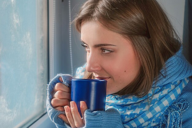 smiling dreaming girl in sweater with the cup near the frozen window in the winter