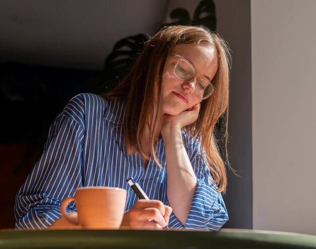 Smiling dreaming woman writing plans in notebook sitting at wood desk in cafe with coffee cup and su...