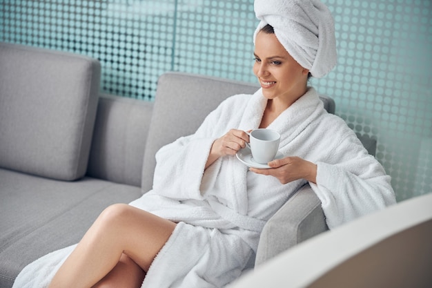 Smiling dreamy lady relaxing with a cup of tea on a couch in a wellness center