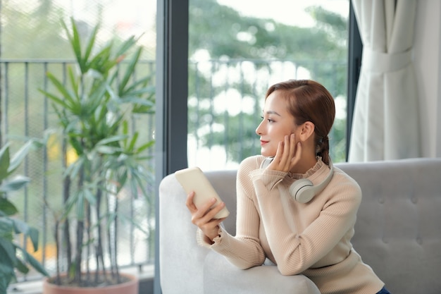 웃고 있는 꿈꾸는 베트남 소녀는 휴대폰을 사용하여 거실 소파에 앉아 멀리서 생각하는 것을 시각화합니다.