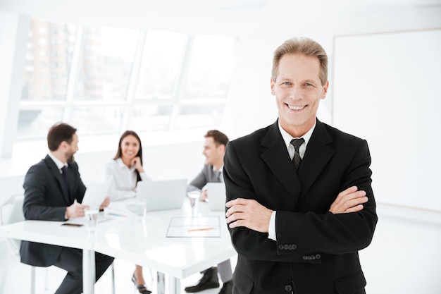 Smiling Elderly Business man standing with arms crossed in office with colleagues