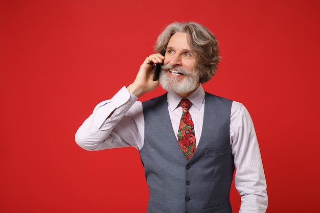 Smiling elderly gray-haired mustache bearded man in classic shirt vest colorful tie posing isolated on red background in studio. People lifestyle concept. Mock up copy space. Talking on mobile phone.