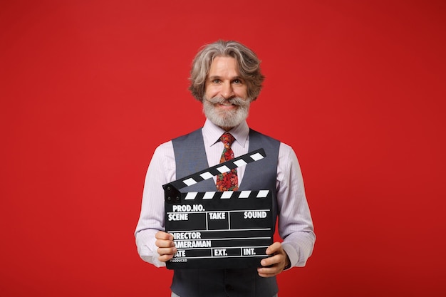 Photo smiling elderly gray-haired mustache bearded man in classic shirt vest and tie isolated on red background. people lifestyle concept. mock up copy space. holding classic black film making clapperboard.