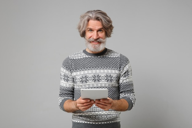 Фото Улыбающийся пожилой седовласый усатый бородатый мужчина в вязаном свитере позирует изолированно на сером фоне стены, студийный портрет. концепция образа жизни людей. скопируйте пространство для копирования. холдинг планшетный компьютер.