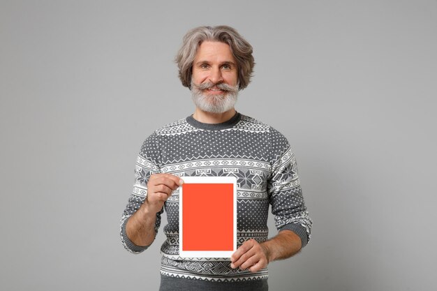 Photo smiling elderly gray-haired mustache bearded man in knitted sweater posing isolated on grey background. people lifestyle concept. mock up copy space. hold tablet pc computer with blank empty screen.