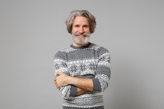 Smiling elderly gray-haired mustache bearded man in knitted\
sweater posing isolated on grey wall background, studio portrait.\
people lifestyle concept. mock up copy space. holding hands\
crossed.