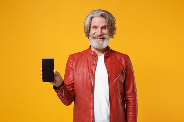 Smiling elderly gray-haired mustache bearded man in red leather jacket isolated on yellow orange background. People lifestyle concept. Mock up copy space. Hold mobile phone with blank empty screen.