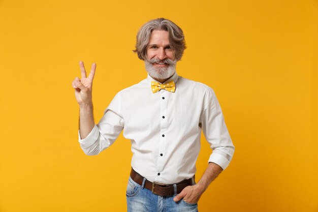 Smiling elderly gray-haired mustache bearded man in white shirt and bow tie posing isolated on yellow orange background in studio. people lifestyle concept. mock up copy space. showing victory sign.