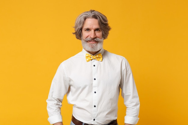 Smiling elderly gray-haired mustache bearded man in white shirt\
and bow tie posing isolated on yellow orange background, studio\
portrait. people sincere emotions lifestyle concept. mock up copy\
space.