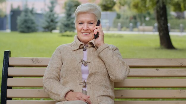 Smiling elderly grayhaired lady with beautiful face sitting on park bench takes phone call woman of retirement age holds smartphone in hand answers by cell has pleasant conversation with relatives