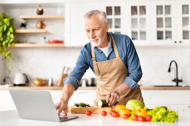Smiling elderly man looking recipe on laptop while cooking food in kitchen
