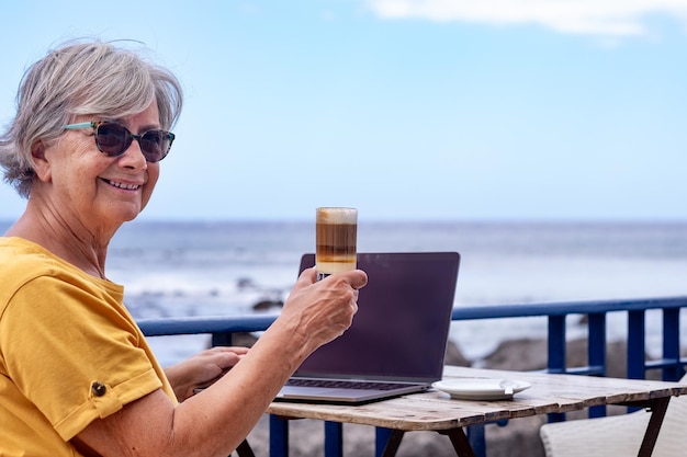 ノートパソコンを使用してカフェのテーブルで屋外に座っている年配の女性の笑顔。アクティブなブロガー遊牧民の労働者