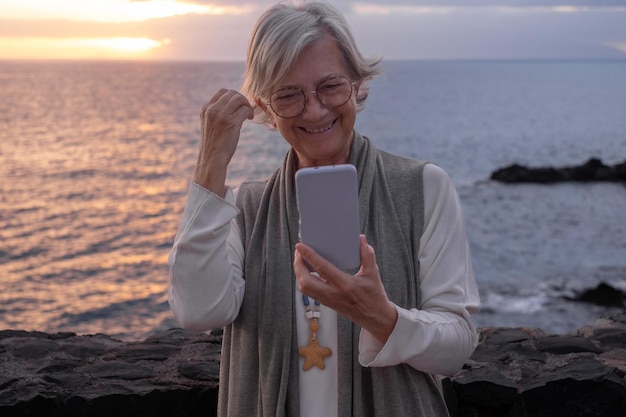 Smiling elderly woman standing in front of the sea looks at her\
mobile phone while the sun sets in the sea peace solitude\
relaxation meditation