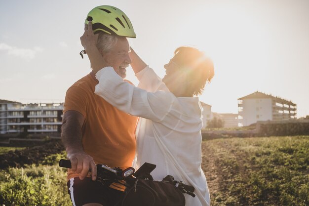 Smiling elderly woman takes care of her cyclist husband by\
putting on his helmet before riding his bike. bright sunset\
light