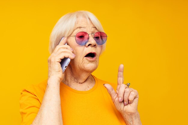 Улыбающаяся пожилая женщина разговаривает по телефону в желтых очках на желтом фоне