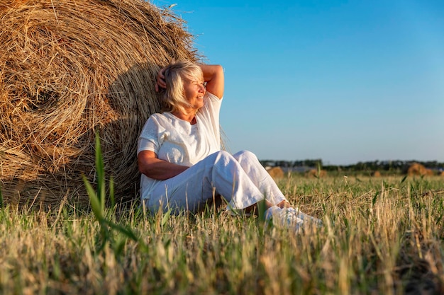 Улыбающаяся пожилая женщина в белом свитере и брюках в поле с рулонами сена на закате. Свобода земледелия, активность на природе и долголетие.