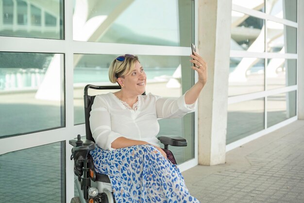 A smiling elegant woman in a wheelchair taking a selfie photo outdoors