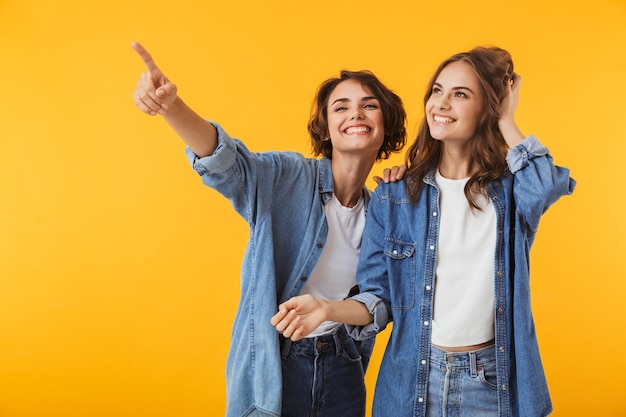 Smiling emotional young women friends posing isolated over yellow wall pointing.