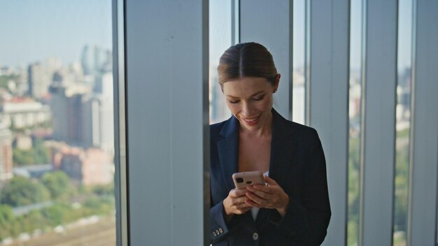 Smiling employee reading smartphone typing message in modern panoramic office