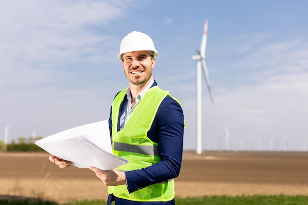 Smiling Engineer Reviewing Plans Near Wind Turbines on a Sunny Day