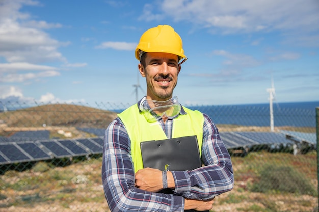 A smiling engineer with folded arms at a solar farm holding a folder