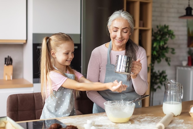 Улыбающаяся европейская маленькая девочка и пожилая женщина в фартуках делают тесто из муки и веселятся в интерьере кухни