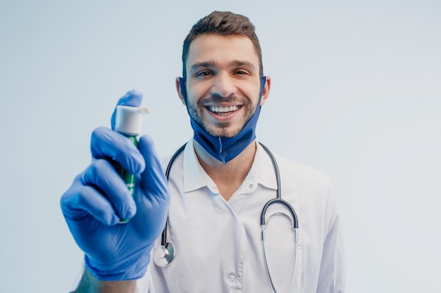 Smiling european male doctor showing inhaler. Young bearded man with stethoscope wearing white coat, protection mask and latex glove. Isolated on gray background with turquoise light. Studio shoot