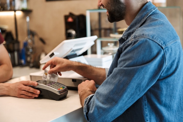 Sorridente uomo europeo che indossa una camicia di jeans pagando la carta di debito nella caffetteria mentre il cameriere tiene il terminale di pagamento